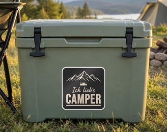 May include: A sage green rectangular cooler with two black rubber latches on the front. A square sticker is centred on the cooler, featuring a white mountain graphic, a small camper icon, and the text Ich lieb's CAMPER in white and beige.