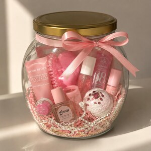 May include: A clear glass jar with a gold lid and a pink ribbon bow containing various pink-themed beauty products, including a Glow Recipe tube, a Gisou bottle, a Sol de Janeiro bottle, a bath bomb, and pink crinkle paper filler inside.