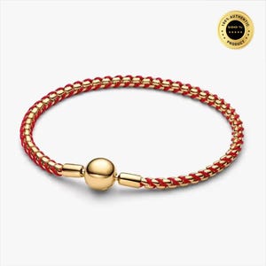 Op de afbeelding: Een armband met een gevlochten rood koord verweven met goudkleurige metalen schakels. Het sieraad bevat een centrale gepolijste goudkleurige bolvormige kraal en een bijpassend cilindervormig sluitingsmechanisme. Een gouden cirkelvormig zegel met de tekst 100% AUTHENTIC is zichtbaar.