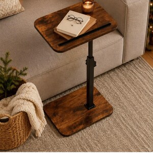 May include: A C-shaped side table with a dark brown wood-grain finish top and base, connected by a black metal adjustable height pole. The tabletop holds a closed book, a pair of black-rimmed glasses, and a small amber glass candle holder.