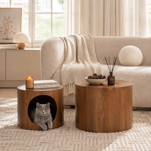 May include: Two cylindrical wooden side tables with a smooth brown finish. The smaller table features a hollow circular opening containing a grey cat. The larger table holds a small bowl of pinecones and a glass reed diffuser with black sticks.
