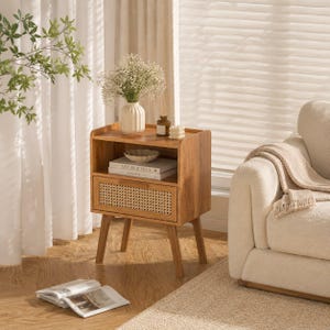 May include: A light brown wooden nightstand with four tapered legs, an open top shelf, and a single drawer featuring a woven cane front panel. The nightstand holds a white ribbed vase with white flowers, a small glass bottle, and two books.