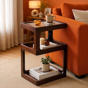 May include: A dark brown wooden side table with three tiers and a fluted glass top. The top tier holds an amber glass candle holder and a ceramic mug. The middle tier features a white candle and a reed diffuser on a book. The bottom tier holds a plant.