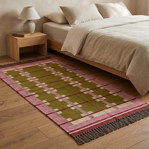 May include: A rectangular woven area rug featuring a repeating geometric pattern in olive green, soft pink, and cream. The rug has dark brown fringe detailing on both short ends and a flat-weave texture, positioned on a light-colored wooden floor.