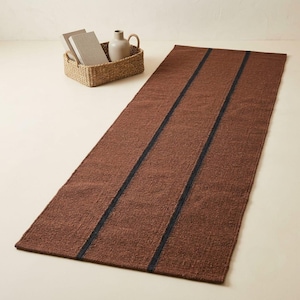 May include: A rectangular brown runner rug featuring two parallel dark charcoal stripes running vertically along its length. The rug has a textured, woven appearance and is positioned on a plain surface near a woven basket containing books.