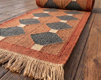 May include: A rectangular woven runner rug featuring a geometric pattern of dark gray diamonds set against a light tan background, bordered by a rust-orange frame. The rug is partially rolled at one end and has a fringed edge at the other.