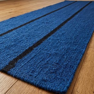 May include: A rectangular runner rug featuring a textured, woven blue surface with two distinct, parallel black stripes running lengthwise across the entire length of the piece. The rug has a thick, knotted construction and a finished edge border.