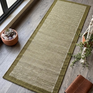 May include: A rectangular runner rug featuring a textured olive green center panel with a lighter, speckled weave, framed by a solid, darker olive green border. The rug has a woven fabric construction with a distinct, clean-edged rectangular shape.