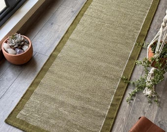 May include: A rectangular runner rug featuring a textured olive green centre panel with a lighter, speckled weave, framed by a solid, darker olive green border. The rug has a woven fabric construction with a distinct, clean-edged rectangular shape.