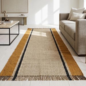 May include: A rectangular woven area rug featuring a natural beige center panel, flanked by wide mustard yellow borders and thin black stripes. The rug has a textured, fibrous appearance and is finished with short, knotted fringe at both ends.