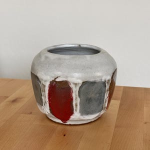 Puede incluir: Un cuenco de cerámica redondo con una abertura ancha, que presenta un esmalte blanco texturizado en la parte superior que gotea sobre una serie de paneles rectangulares en tonos alternos de rojo intenso, gris oscuro y marrón en el cuerpo inferior.