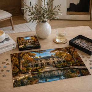 May include: A jigsaw puzzle featuring a large mansion surrounded by autumn trees with golden foliage and a reflecting pool. The puzzle box shows the same image, and a black tray contains loose puzzle pieces scattered on a light wooden table.