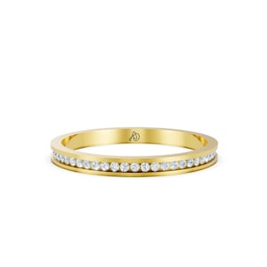May include: A yellow gold band ring featuring a channel setting with a continuous row of small, round, clear, sparkling stones embedded along the center. The interior of the band displays a small, black, stylized logo mark near the top edge.