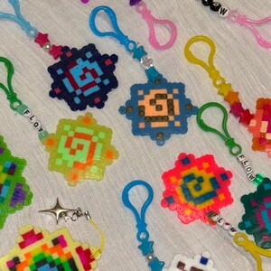 Puede incluir: Una colección de coloridos llaveros de pixel art hechos de cuentas de plástico fundido. Cada llavero presenta un diseño cuadrado o en forma de estrella con patrones geométricos, unido a un mosquetón de plástico con cuentas en forma de estrella y algunos deletrean la palabra FLOW.