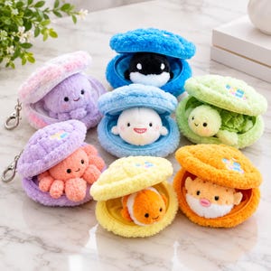 May include: A collection of six plush clam-shaped pouches, each containing a small stuffed sea creature. The set includes a purple octopus, a black and white orca, a white beluga, a green turtle, an orange octopus, a clownfish, and a pufferfish.