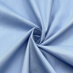 Puede incluir: Una tela de color azul claro sólido con una textura suave y mate. El material está recogido en el centro, creando una serie de pliegues radiantes y sombras profundas que resaltan el color consistente y el tejido fino de la superficie.