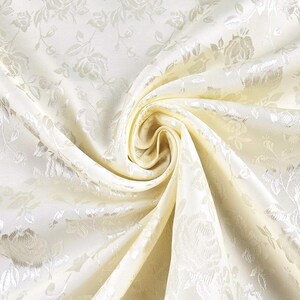 Puede incluir: Una tela de color crema con un patrón de rosas en jacquard tonal. El material está recogido en el centro, creando pliegues y drapeados suaves que resaltan el brillo del diseño floral tejido en la superficie del textil.