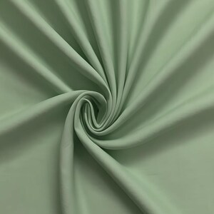 Peut inclure: Un tissu vert sauge clair uni avec une texture lisse et mate. Le matériau est rassemblé au centre, créant des plis doux et rayonnants ainsi que des ombres profondes qui soulignent la couleur uniforme et la surface régulière du textile.