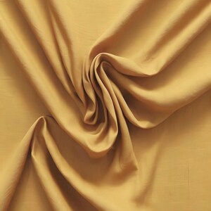 Könnte beinhalten: Ein einfarbiger senfgelber Stoff mit einer glatten, matten Textur. Das Material ist in weichen, fließenden Falten drapiert, die natürliche Schatten und Glanzlichter auf der Oberfläche erzeugen und die gleichmäßige Farbe sowie das feine Gewebe betonen.