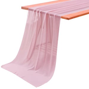 May include: A long, sheer, light pink table runner made of a lightweight, semi-transparent fabric. The runner is draped over a wooden surface, with one end extending downward, showcasing the soft, flowing folds and the delicate, airy texture.