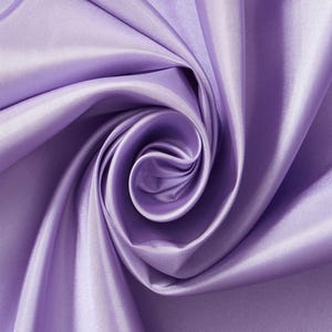 May include: A length of solid light purple fabric with a smooth, lustrous finish. The material is arranged in a central spiral swirl, creating deep, flowing folds and shadows that highlight the reflective, satin-like texture of the textile.