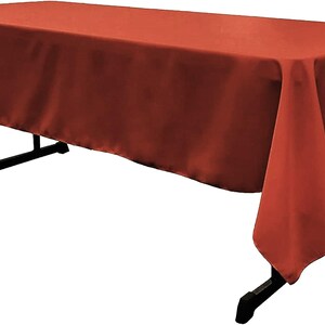 May include: A rectangular tablecloth in a solid burnt orange color draped over a black folding table. The fabric has a smooth, matte finish and hangs evenly over the edges of the table, covering the top and sides while leaving the legs visible.