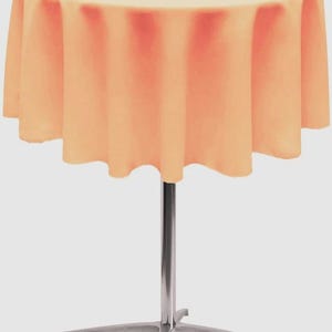 May include: A round cocktail table featuring a solid peach-colored fabric tablecloth that drapes evenly around the edges. The table is supported by a central silver-toned metal pole and a four-pronged base made of the same metallic material.