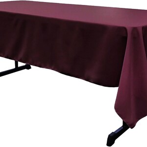 May include: A rectangular burgundy tablecloth draped over a black folding table. The fabric has a smooth, solid-colored finish that covers the top and hangs down over the sides, creating clean, straight edges and soft folds at the corners.