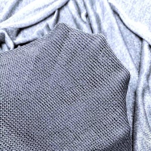 Può includere: Un tessuto grigio chiaro con una distinta trama a rete a maglia aperta. Il materiale è semi-trasparente e presenta un motivo fine a griglia su tutta la superficie. Il tessuto è drappeggiato in morbide pieghe, evidenziando la costruzione leggera.
