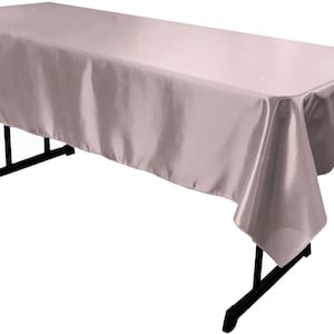 Pode incluir: Uma mesa retangular coberta com uma toalha de cetim rosa claro sólido. O tecido tem um acabamento liso e reflexivo que cai sobre as bordas da mesa dobrável preta, que possui uma estrutura de metal com quatro pernas e barras de suporte.