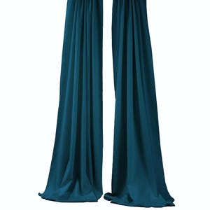 May include: Two long, floor-length teal curtains hanging from a dark brown horizontal rod. The fabric features vertical folds and drapes downward, creating a pleated effect. The curtains are opaque and solid in color, extending from the top rod to the bottom edge.