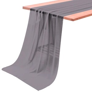 Puede incluir: Un camino de mesa largo y rectangular hecho de una tela gris semitransparente. El camino está colocado sobre el borde de una superficie de madera, creando pliegues verticales suaves que se extienden desde el borde superior hasta el dobladillo.