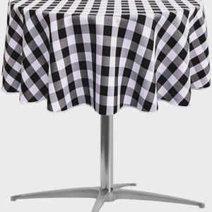 May include: A round table covered with a black and white checkered tablecloth. The fabric features a repeating square pattern that drapes over the edges of the circular tabletop, supported by a central silver metal pedestal base with four legs.