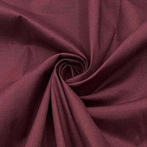 Puede incluir: Una tela de color burdeos oscuro sólido con una textura suave y mate. El material está recogido en el centro, creando pliegues profundos y fluidos y sombras que irradian hacia afuera, resaltando el color rojo púrpura profundo del textil.