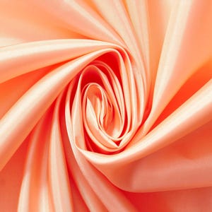 May include: A smooth, peach-colored fabric with a subtle sheen, arranged in a swirling, circular pattern that creates deep, flowing folds and highlights across the surface, emphasizing the soft texture and light-reflecting quality of the material.