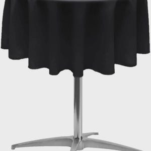 May include: A round cocktail table featuring a solid black fabric tablecloth that drapes over the edges. The table is supported by a central silver-colored metal pedestal column and a four-pronged silver metal base with flat feet.
