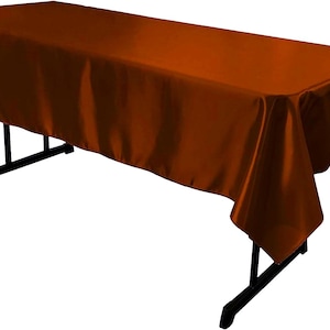May include: A rectangular tablecloth in a solid burnt orange color with a smooth, satin-like finish. The fabric drapes over the edges of a black folding table, covering the top surface and extending down the sides with soft, flowing folds.