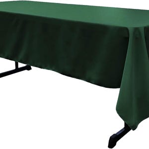 May include: A rectangular dark green tablecloth draped over a black folding table. The fabric has a smooth, solid-colored surface that covers the top and hangs down over the sides, while the black metal legs of the table are visible underneath.