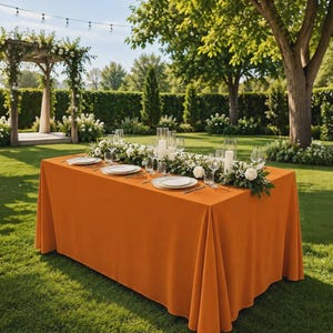 May include: A rectangular table covered with a solid orange tablecloth, set with white plates, clear glassware, and tall cylindrical glass candle holders. A long floral centerpiece of white roses and greenery runs down the center of the table.