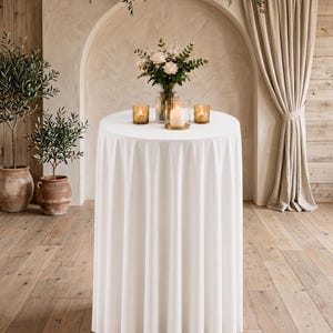 Può includere: Un tavolo da cocktail alto e rotondo coperto da una tovaglia bianca che arriva fino a terra con pieghe verticali. Sul piano del tavolo si trova un vaso di vetro trasparente con rose bianche e verde, affiancato da tre piccoli portacandele.