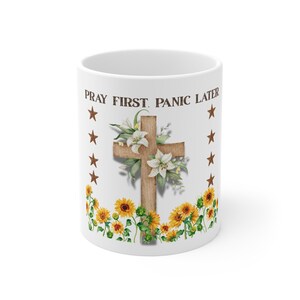May include: A white ceramic mug featuring the text PRAY FIRST. PANIC LATER. in dark brown letters. The design includes a central wooden cross adorned with white lilies, surrounded by yellow sunflowers at the base and brown stars on the sides.