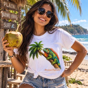 Op de afbeelding: Een wit T-shirt met korte mouwen en een afbeelding van een groene leguaan op een palmboomstam, met de tekst Ancianos Tropicales in het oranje onder de illustratie. Het shirt wordt gedragen met een blauwe spijkerbroek en een zonnebril.