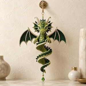 May include: A hanging crochet dragon ornament in shades of light and dark green. The creature features detailed wings, horns, and a long, spiraling tail. It is suspended from a gold-toned wall hook by a thin chain with a small green crystal pendant.
