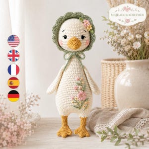 May include: A cream-colored crocheted duck toy featuring an orange beak and feet, wearing a sage green scalloped bonnet with small pink and yellow flowers. The duck's body is decorated with embroidered pink and yellow floral sprigs on the front.