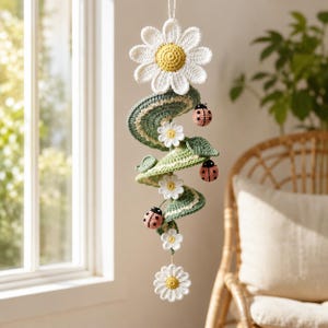 Könnte beinhalten: Ein hängendes Häkel-Mobile mit einer großen weißen Margerite mit gelber Mitte an der Spitze, gefolgt von einer spiralförmigen grünen Rankenstruktur, die mit kleineren weißen Margeriten und drei kleinen rosa Marienkäfern mit schwarzen Punkten verziert ist.
