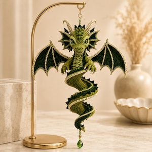 May include: A green crocheted dragon ornament hanging from a gold metal stand. The dragon features detailed wings, a long coiled tail, and small horns. A green crystal bead hangs from the bottom of the tail, and the dragon has large eyes.