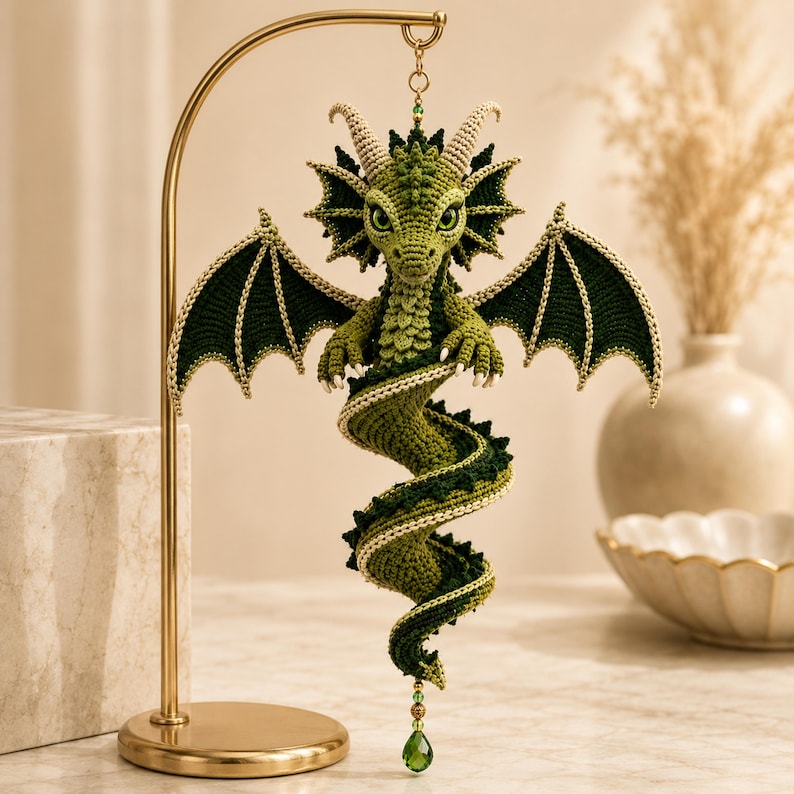 May include: A green crocheted dragon ornament hanging from a gold metal stand. The dragon features detailed wings, a long coiled tail, and small horns. A green crystal bead hangs from the bottom of the tail, and the dragon has large eyes.