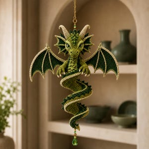 May include: A hanging crochet dragon ornament in shades of olive and forest green. The creature features textured scales, two outstretched wings with cream-colored edges, small horns, and a long, spiraling tail ending in a green crystal bead.