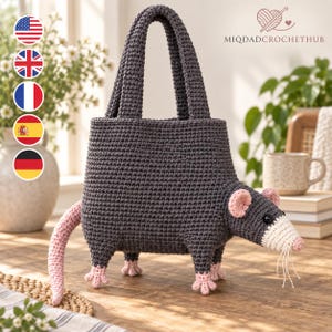 May include: A dark grey crocheted bag shaped like an opossum with a cream-coloured snout, pink ears, pink feet, and a long pink tail. The bag features two top handles and black button eyes. The text MIQDADCROCHETHUB is printed in the upper right corner.