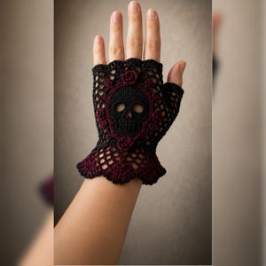 May include: A fingerless crochet glove featuring a black skull motif centred within a dark red ornate frame. The glove is crafted from black and dark red yarn in an intricate openwork lace pattern, with a scalloped edge at the wrist area.